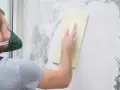 Mężczyzna w masce ochronnej nakłada tynk mozaikowy na ścianę za pomocą kielni.