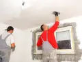 Dwóch budowlańców tynkuje sufit i ściany w pomieszczeniu.