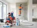 Dwóch budowlańców tynkuje ściany w nowym budynku, używając maszyny tynkarskiej.