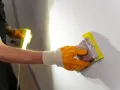 Ręka w pomarańczowej rękawicy szlifuje ścianę, przygotowując ją pod malowanie. Drobny pył unosi się w powietrzu, a światło lampy podkreśla nierówności, które znikną po nałożeniu gładzi.