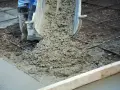 Wylewanie świeżego betonu na zbrojenie. Trwa naprawa betonu, widać buty pracownika i metalowe pręty.