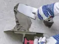 Naprawa betonu na zewnątrz. Ręce w rękawicach nakładają zaprawę kielnią na uszkodzony fragment ściany.