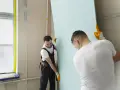 Dwóch budowlańców w rękawiczkach montuje dużą, jasnoniebieską płytę gipsowo-kartonową. Prace przy łączeniu płyt gk postępują sprawnie.