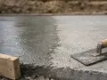 Świeżo wylany, mokry beton czeka na wykończenie. Narzędzia budowlane, jak kielnia i drewniany krawężnik, sugerują szybki postęp prac.