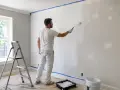 Mężczyzna w białym ubraniu wykonuje gruntowanie ścian wałkiem. Obok drabina i wiadro z farbą.