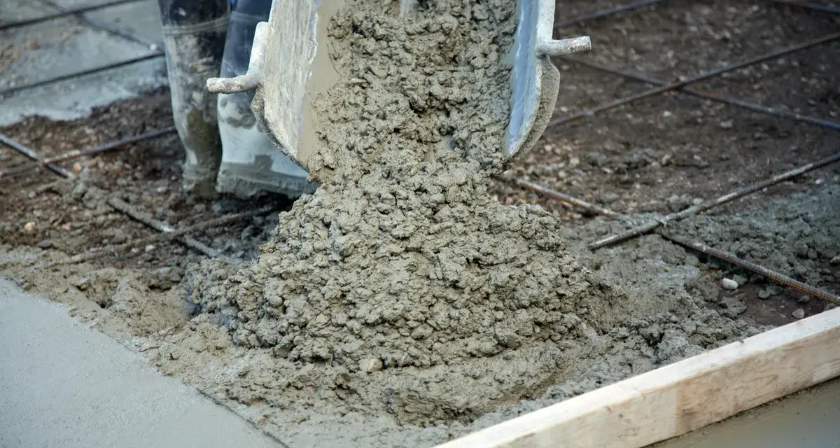 Wylewanie świeżego betonu na zbrojenie. Trwa naprawa betonu, widać buty pracownika i metalowe pręty.