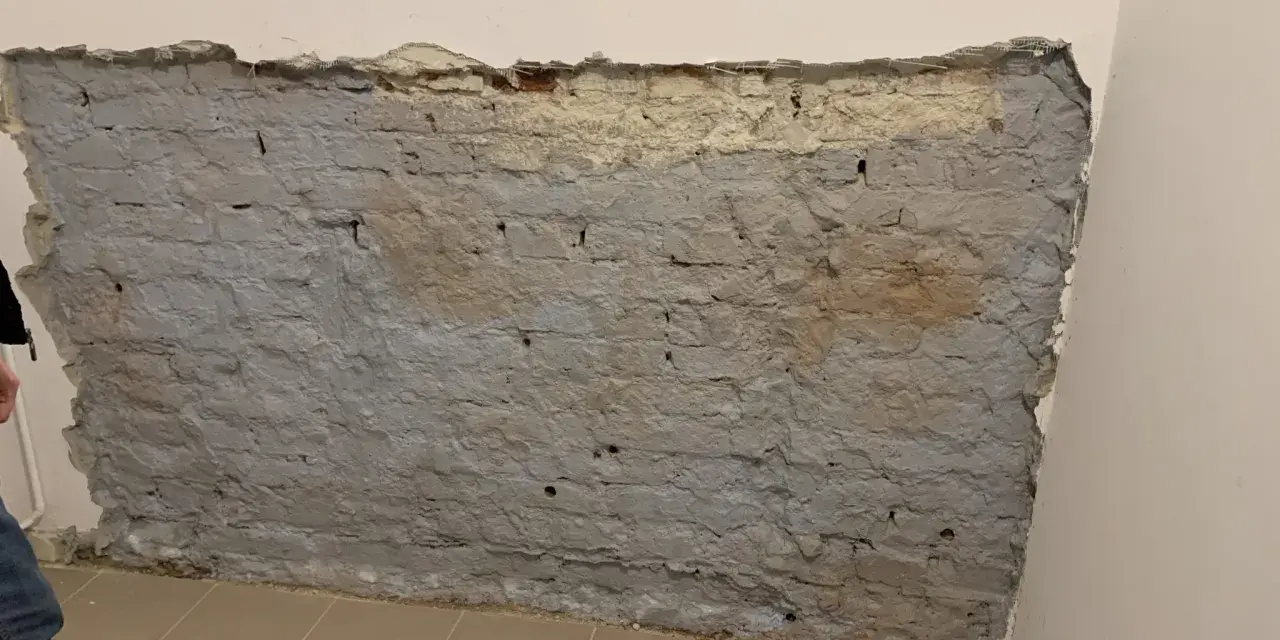 Fragment odsłoniętej, starej ceglanej ściany z szarym tynkiem i fragmentami żółtego podkładu.