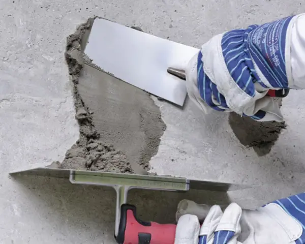 Naprawa betonu na zewnątrz. Ręce w rękawicach nakładają zaprawę kielnią na uszkodzony fragment ściany.