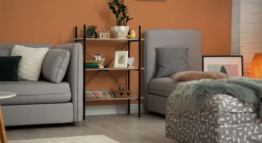 Przytulny salon z sofą i fotelem, ściany w ciepłym, **kolorze ceglastym**, regał z dekoracjami.