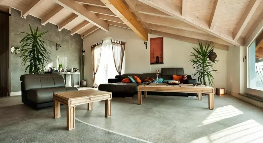 Nowoczesny salon z drewnianymi belkami i szarymi podłogami epoksydowymi. Wygodne kanapy, drewniane stoliki i dużo zieleni tworzą przytulną atmosferę.