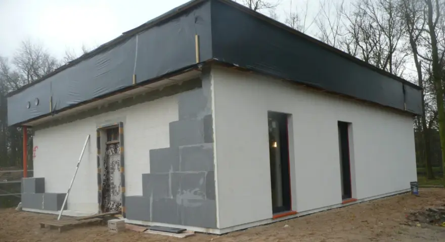 Budowa domu ze styropianu w toku. Ściany z bloczków, częściowo otynkowane, dach przykryty czarną folią.