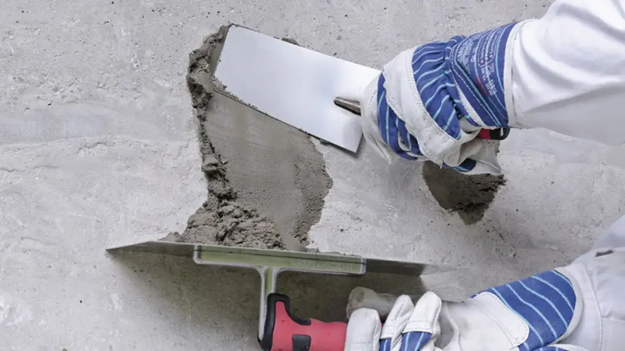Naprawa betonu na zewnątrz. Ręce w rękawicach nakładają zaprawę tynkarską na uszkodzony fragment ściany za pomocą kielni.