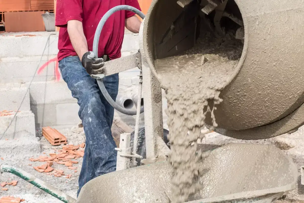 Robotnik miesza chudy beton, dbając o właściwe proporcje składników. Mieszanka spływa z betoniarki do taczki.