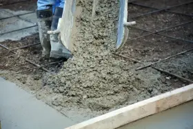 Wylewanie świeżego betonu na zbrojenie. Trwa naprawa betonu, widać buty pracownika i metalowe pręty.