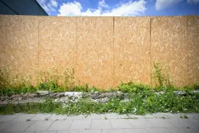 Płyta OSB na zewnątrz, otoczona dziką zielenią i fragmentami gruzu, tworzy tymczasowe ogrodzenie.