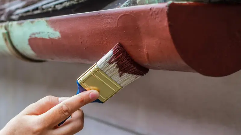 Ręka maluje pędzlem rynienkę aluminiową na brązowo. Odpowiedź na pytanie: czym pomalować aluminium.